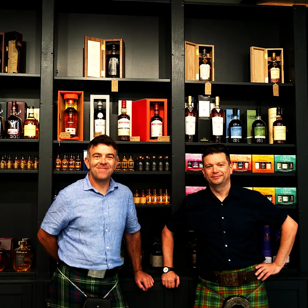 Two of the Wild Woven team in kilts in front of the whisky wall