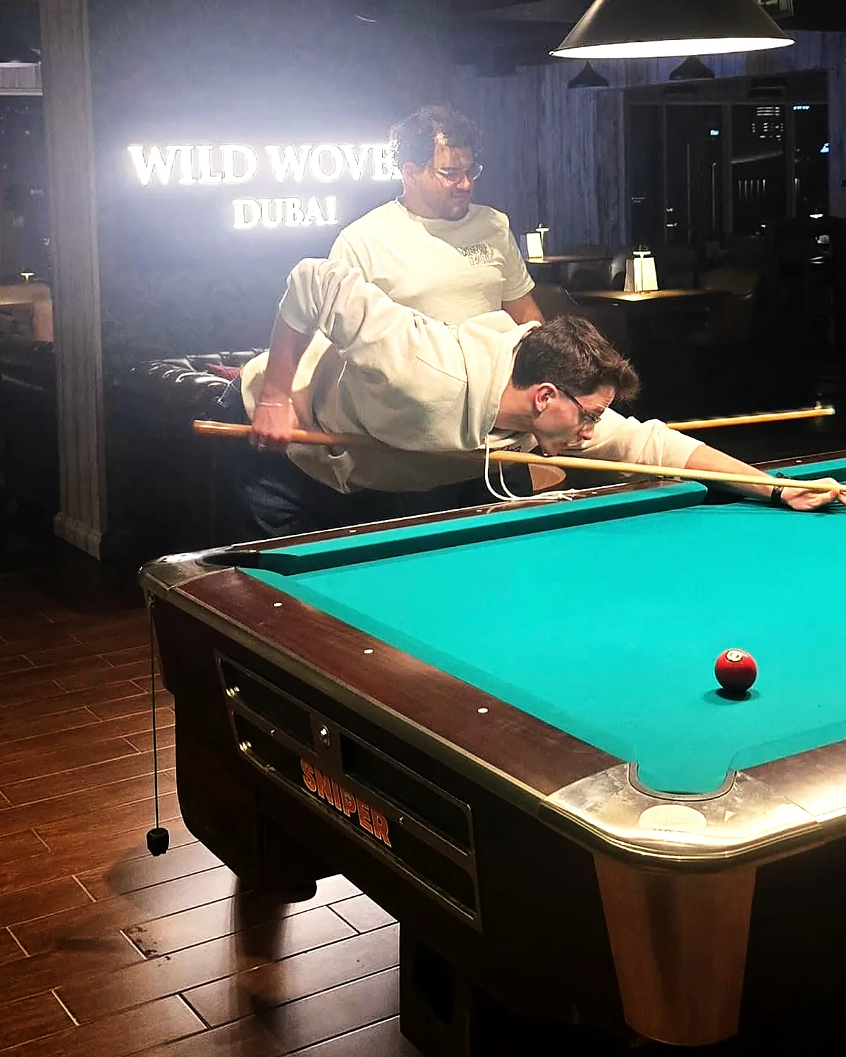 Two guests playing billiards beneath the illuminated Wild Woven Dubai sign